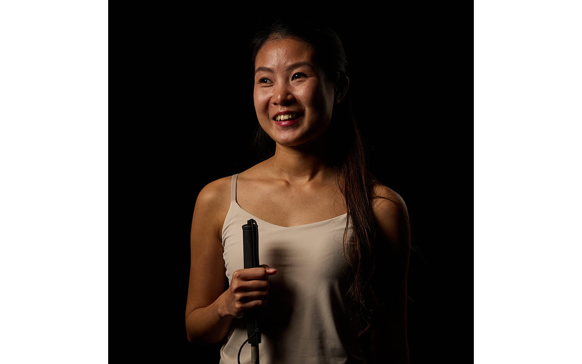 Claire Teo, the author, stands against a dark background, smiling softly. She holds a white cane upright in one hand, facing slightly to the side.