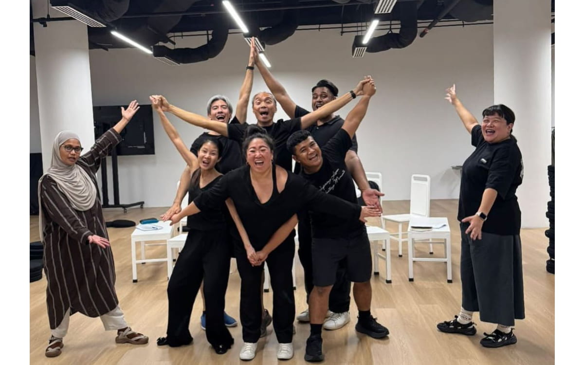 In rehearsal, two actors stand at the far left and right with arms open wide. In the centre, seven actors including Claire, cluster together in a silly, star-like formation, reaching their arms outward and upward while smiling widely.