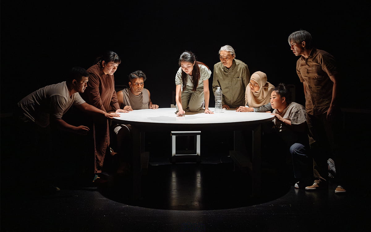 Water has been spilled on a round table. Seven actors gather around, all leaning in. At the centre, Claire kneels on the table, reaching towards her reflection.
