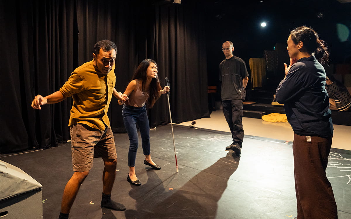 Four people on a stage in rehearsal. On the left, an actor leans forward tensely while Claire, beside him, grips a white cane and appears to shout. Yvan, the director, stands to the right watching, while the other actor faces them while on the phone.