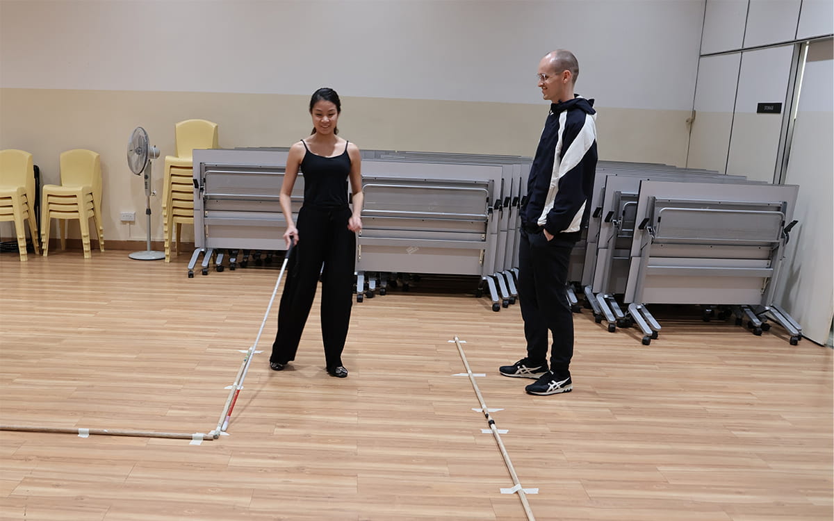 Claire and Yvan in a rehearsal space with a wooden floor. On the left, Claire uses her cane to trace along taped, raised lines forming a square on the floor. On the right, Yvan, stands nearby watching her.