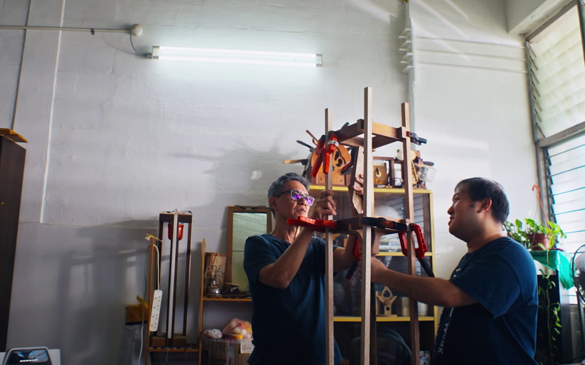 Two men, one in his 60s and the other in his 30s, having a conversation in an artist studio. Their hands are placed on a structure made out of thin wooden poles and planks, held together by red clamps. 