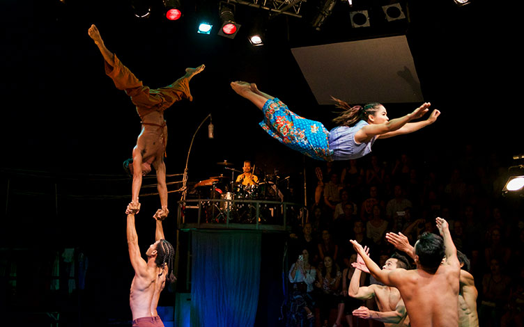 A female circus performer soars through the air as her fellow performers stand ready to catch her.