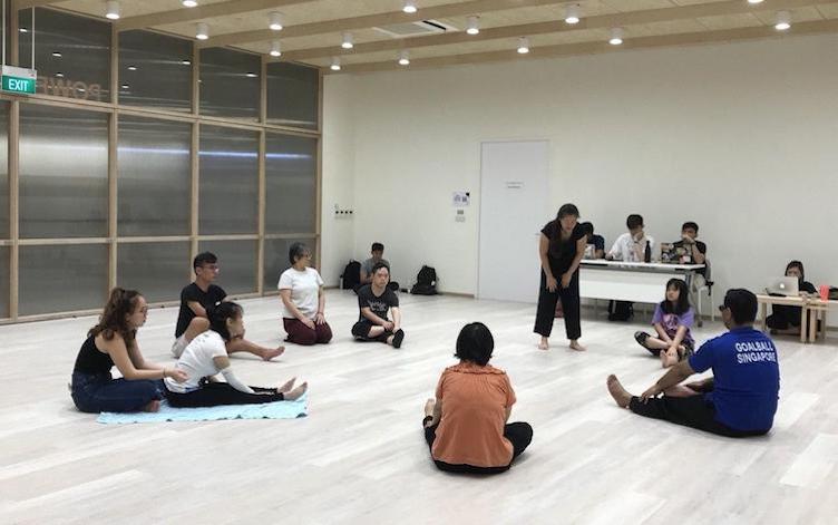 Director, Jing Hong and the cast during warm-ups. Cast is seated on the floor.