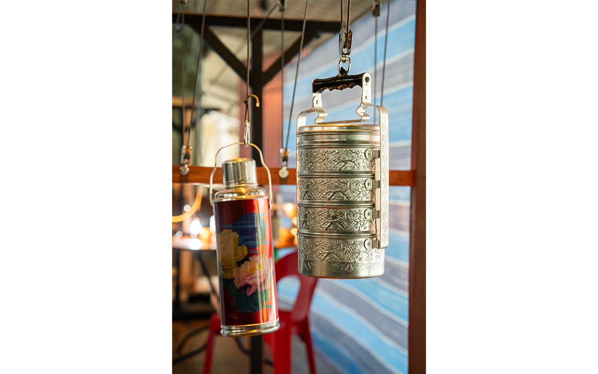 A tingkat and a colourful metal flask hang in front of blue striped tarpaulin.