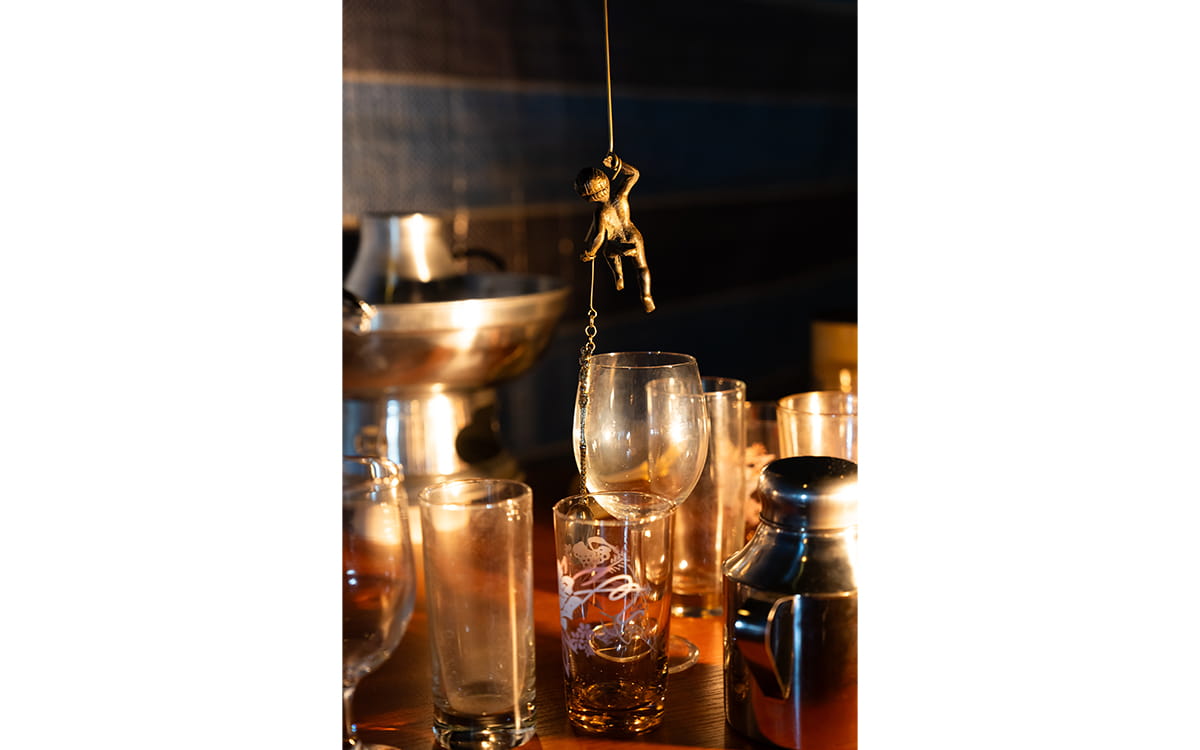 A small wooden figurine of a man wearing a helmet climbing a rope suspended above a glass cup, surrounded by other empty glasses and dinnerware on a dimly lit table.