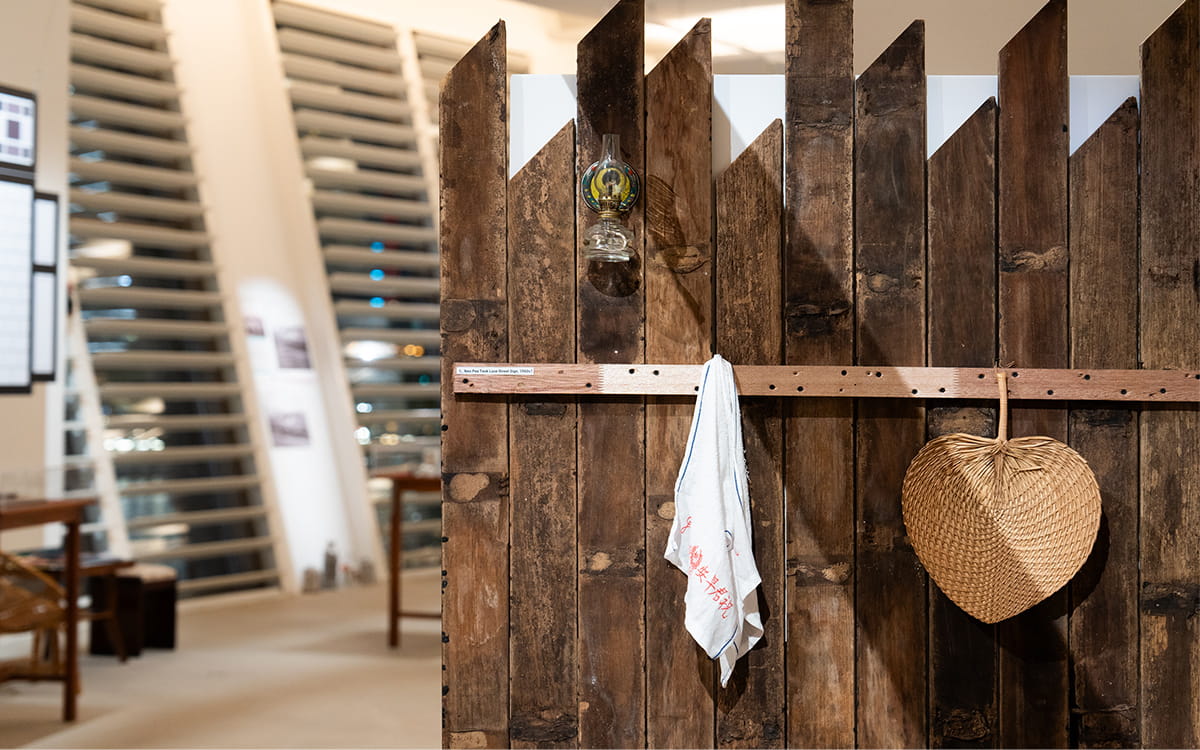 Close-up of a wooden fence installation with a small kerosene lamp, Good Morning towel, and rattan fan hanging from hooks.