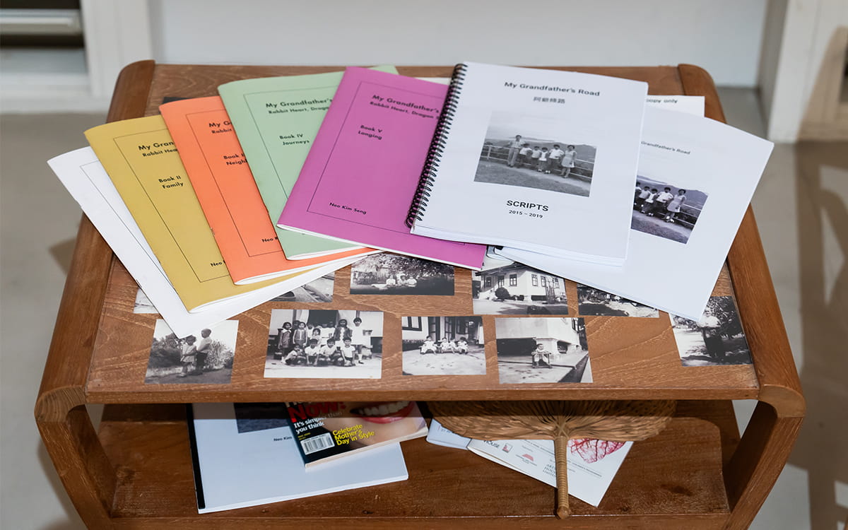On a small wooden side table sits a collection of colourful books and scripts of My Grandfather's Story, arranged on top of some old black and white childhood photos.